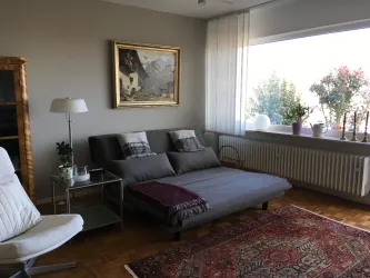 Ein helles Wohnzimmer mit einem bequemen Schlafsofa und einem Fenster, das auf Pflanzen blickt. An der Wand hängt ein Gemälde, und im Raum stehen ein Sessel sowie ein Tisch.