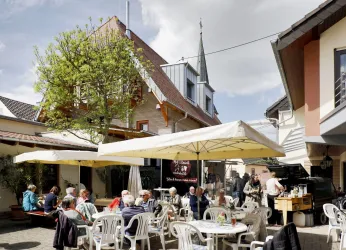 Ein gemütliches Café im Freien mit vielen Tischen und Stühlen. Menschen genießen die Sonne und die entspannte Atmosphäre.