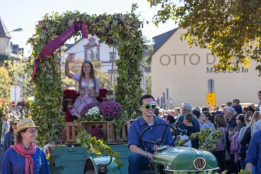 Eine festliche Parade mit einem geschmückten Traktor. Eine junge Frau sitzt auf dem Wagen und wird von einer Menschenmenge umgeben.