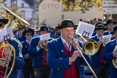 Eine Blaskapelle in traditionellen Anzügen spielt während einer Parade. Die Musiker tragen Hüte und sind in festlicher Stimmung.