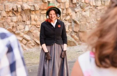 Eine lächelnde Frau trägt eine traditionelle Tracht und einen Hut. Sie steht vor einer Felswand und wirkt fröhlich.