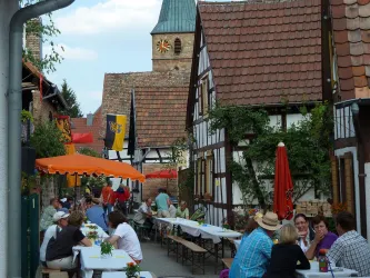 Eine malerische Gasse mit Fachwerkhäusern und Menschen, die an Tischen sitzen. Bunte Sonnenschirme sorgen für eine einladende Atmosphäre.