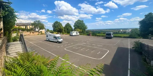 Ein großer, leerer Parkplatz umgeben von Bäumen und grünem Gelände. Der Himmel ist blau mit einigen weißen Wolken.