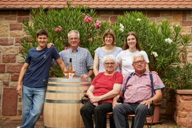 Eine Familiengruppe steht vor einem Weinfass und blühenden Pflanzen. Alle lächeln und wirken freundlich.
