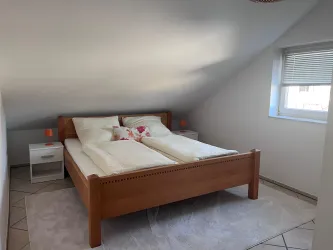 Ein gemütliches Schlafzimmer mit einem großen Holzbett und zwei Nachttischen. Das Zimmer hat schrägen Wände und ein Fenster, das natürliches Licht hereinlässt.