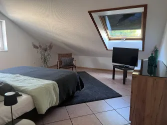 Ein gemütliches Schlafzimmer mit einem großen Bett und einem Fernseher. Der Raum hat ein modernes Design und ein Fenster mit Tageslicht.