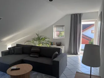 Ein helles Wohnzimmer mit einem grauen Sofa und einem runden Holztisch. Durch ein Fenster scheint Tageslicht und eine Pflanze steht neben dem Sofa.