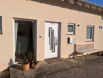 Ein moderner Hauseingang mit einer weißen Tür und einem kleinen Vorgarten. Neben der Tür steht eine Bank und es gibt Töpfe mit Pflanzen.