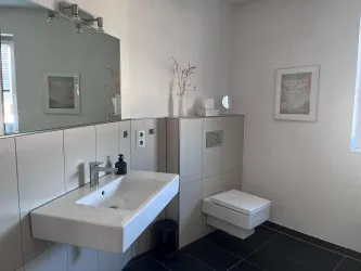 Ein modernes Badezimmer mit einer weißen Wand- und Bodenfliese. Der Raum enthält ein Waschbecken, eine Toilette und einen großen Spiegel.