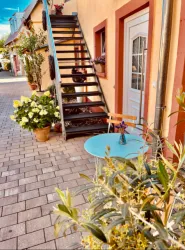 Ein schöner Außenbereich mit einer Metalltreppe und blühenden Pflanzen. Ein runder blauer Tisch steht vor einer Tür, umgeben von weiteren Pflanzen.