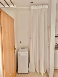 Ein kleiner Raum mit einer Waschmaschine und einem weißen Vorhang. Die Wand ist hell und es gibt eine Holztür auf der linken Seite.