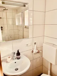 Ein modernes Badezimmer mit weißen Fliesen und einem runden Waschbecken. Zu sehen sind ein Spiegel, ein Handtuchhalter und eine Dusche.