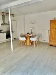 Ein helles Esszimmer mit einem runden Tisch und modernen Stühlen. Im Hintergrund befindet sich eine offene Küche und ein Holzschrank.