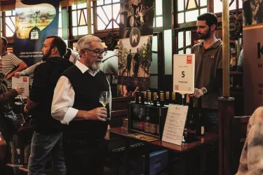 Eine Weinmesse mit mehreren Ständen und Menschen, die Wein verkosten. Ein älterer Mann hält ein Weinglas und spricht mit einem Standbetreuer.
