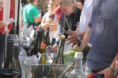 Ein Marktstand mit einer Auswahl an Weinen und Getränken. Menschen stehen in der Nähe und interagieren miteinander.