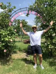 Ein fröhlicher Mann steht unter einem gebogenen Tor mit dem Schriftzug „Mendel-Lernplatz“. Um ihn herum sind Blumen und grüne Bäume zu sehen.