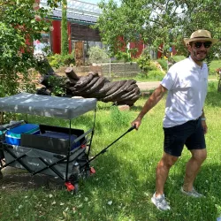 Ein Mann zieht einen Wagen durch eine grüne Wiese. Im Hintergrund sind Pflanzen und eine Skulptur zu sehen.