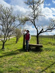 Ein Mann steht lächelnd unter einem Baum in einer grünen Landschaft. Im Hintergrund sind Weinreben und ein blauer Himmel mit Wolken zu sehen.