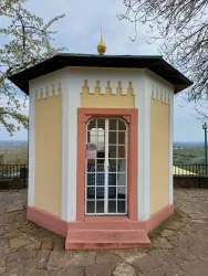 Ein kleiner Pavillon mit einer oktogonalen Form, bemalt in Gelb und Rosa. Er steht in einer aussichtsreichen Lage, umgeben von Bäumen.