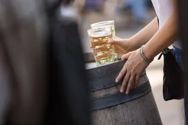 Eine Person hält zwei Gläser mit Getränken. Sie steht neben einem hölzernen Fass.