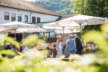 Eine belebte Terrasse mit vielen Gästen unter Sonnenschirmen. Im Hintergrund sind Gebäude und die grüne Landschaft sichtbar.