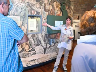 Eine Museumsführerin erklärt den Besuchern die Ausstellung. Im Hintergrund ist eine historische Wandmalerei mit Menschen zu sehen.