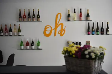 Eine Wand mit verschiedenen Weinflaschen in einer modernen Umgebung. Im Vordergrund steht ein Korb mit bunten Blumen.