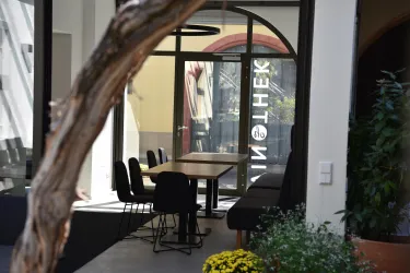 Ein moderner Innenraum mit einem großen Tisch und Stühlen. Im Hintergrund ist eine Fensterfront zu sehen, die auf einen hellen Außenbereich blickt.