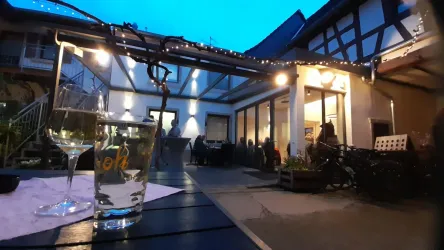 Eine gemütliche Terrasse bei Dämmerung mit warmem Licht. Gläser stehen auf einem Tisch und einige Gäste sind im Hintergrund zu sehen.