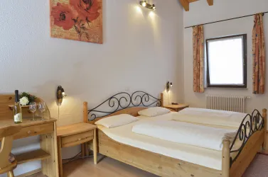 Ein gemütliches Schlafzimmer mit einem Doppelbett und Holzmöbeln. Eine Flasche Wein und Gläser stehen auf einem Tisch, die Fenster lassen viel Tageslicht hinein.