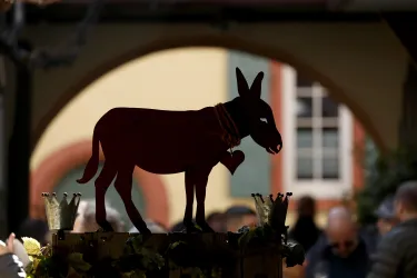 Eine Silhouette eines Esels steht auf einem Tisch. Im Hintergrund sind unscharfe Menschen und eine farbige Wand zu sehen.