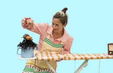 Eine Frau spielt mit einer Puppenfigur, die einen Vogel darstellt. Sie sitzt an einem Tisch, auf dem ein Bügeltuch liegt.