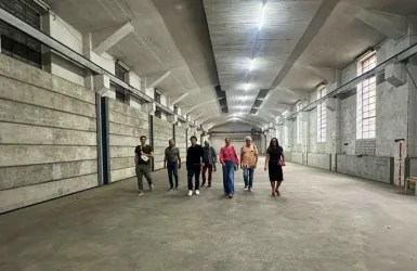 Eine Gruppe von Menschen geht durch einen großen, leeren Raum mit hohen Decken und Betonwänden. Das Licht kommt von den Fenstern und den Deckenlampen.