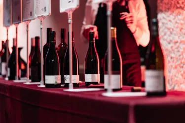 Eine lange Theke mit verschiedenen Flaschen Wein steht im Vordergrund. Im Hintergrund ist eine Person mit einem weißen Hemd zu sehen, die sich um die Weine kümmert.