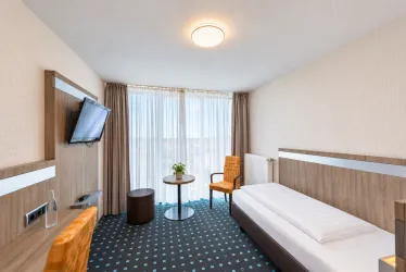 Ein modernes Hotelzimmer mit einem Einzelbett, einem Tisch und einer gemütlichen Ecke. Große Fenster lassen viel Tageslicht hereinzutreten.