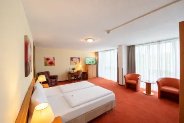 Ein modernes Hotelzimmer mit zwei Betten und orangefarbenem Teppich. Es gibt einen Tisch, Stühle und große Fenster, die viel Tageslicht hereinlassen.
