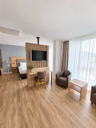 Ein modernes Hotelzimmer mit Holzfußboden und großen Fenstern. Es gibt einen Schreibtisch, zwei Stühle und einen Fernseher.