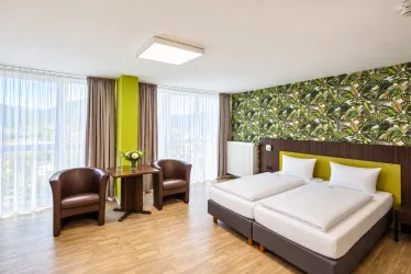 Ein modernes Hotelzimmer mit zwei Betten und einem gemütlichen Sitzbereich. Große Fenster sorgen für viel Tageslicht und eine ansprechende Atmosphäre.