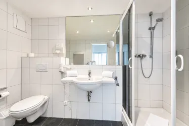 Ein modernes Badezimmer mit weißen Fliesen und klaren Linien. Es enthält eine Dusche, ein Waschbecken und eine Toilette.