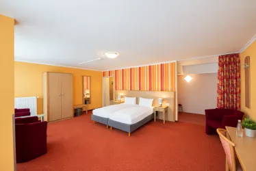 Ein gemütliches Zimmer mit zwei Einzelbetten und rotem Teppich. Die Wände sind in warmen Farben gestaltet, und es gibt Platz für Sitzgelegenheiten.