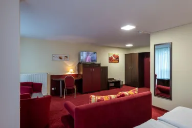 Ein gemütliches Hotelzimmer mit roten Teppichen und einer Sitzecke. Es gibt einen Fernseher, einen Schreibtisch und einen großen Spiegel.
