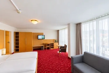 Ein modernes Hotelzimmer mit zwei Betten, einer Couch und einem Tisch. Große Fenster sorgen für viel natürliches Licht und eine gemütliche Atmosphäre.
