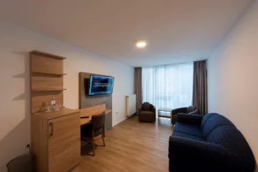 Ein modernes Wohnzimmer mit einem blauen Sofa, einem Holztisch und einem Fernseher an der Wand. Große Fenster lassen viel Licht herein und bieten einen Blick nach draußen.