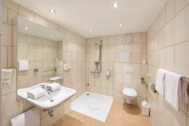 Ein modernes Badezimmer mit hellen Fliesen und einer großzügigen Dusche. Es gibt ein Waschbecken, eine Toilette und frische Handtücher.
