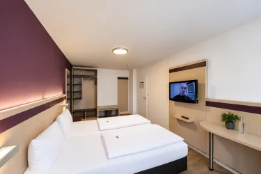 Ein modernes Hotelzimmer mit zwei Einzelbetten und einem Fernseher an der Wand. Die Wände sind in sanften Farben gehalten, und es gibt einen kleinen Tisch mit einer Pflanze.