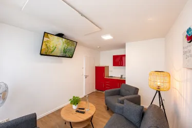 Ein modernes Wohnzimmer mit grauen Sofas und einem Holztisch. Die Küche ist in einer auffälligen roten Farbe und ein großer Fernseher hängt an der Wand.