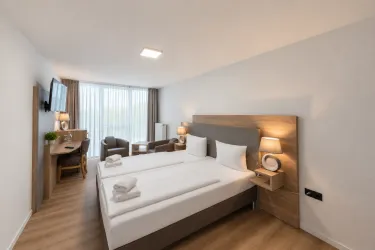 Ein modernes Hotelzimmer mit zwei Betten und neutraler Farbgestaltung. Es gibt einen Schreibtisch, zwei Stühle und große Fenster, die natürliches Licht hereinlassen.