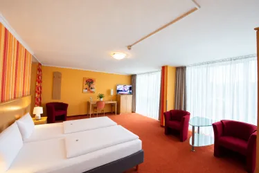 Ein modernes Hotelzimmer mit zwei Einzelbetten und rot-beigen Wänden. Es gibt einen Tisch, zwei rote Stühle und große Fenster, die viel Tageslicht hereinlassen.