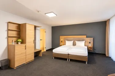 Ein modernes Hotelzimmer mit zwei Einzelbetten und einfachem Holzmobiliar. Die Wände sind in neutralen Farben gehalten und es gibt viel Tageslicht.