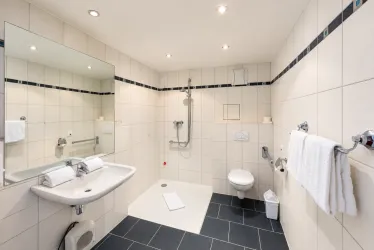 Ein modernes Badezimmer mit weißer Fliesenwand und schwarzen Fliesenboden. Es gibt eine Dusche, ein Waschbecken und eine Toilette, alles sauber und gut beleuchtet.